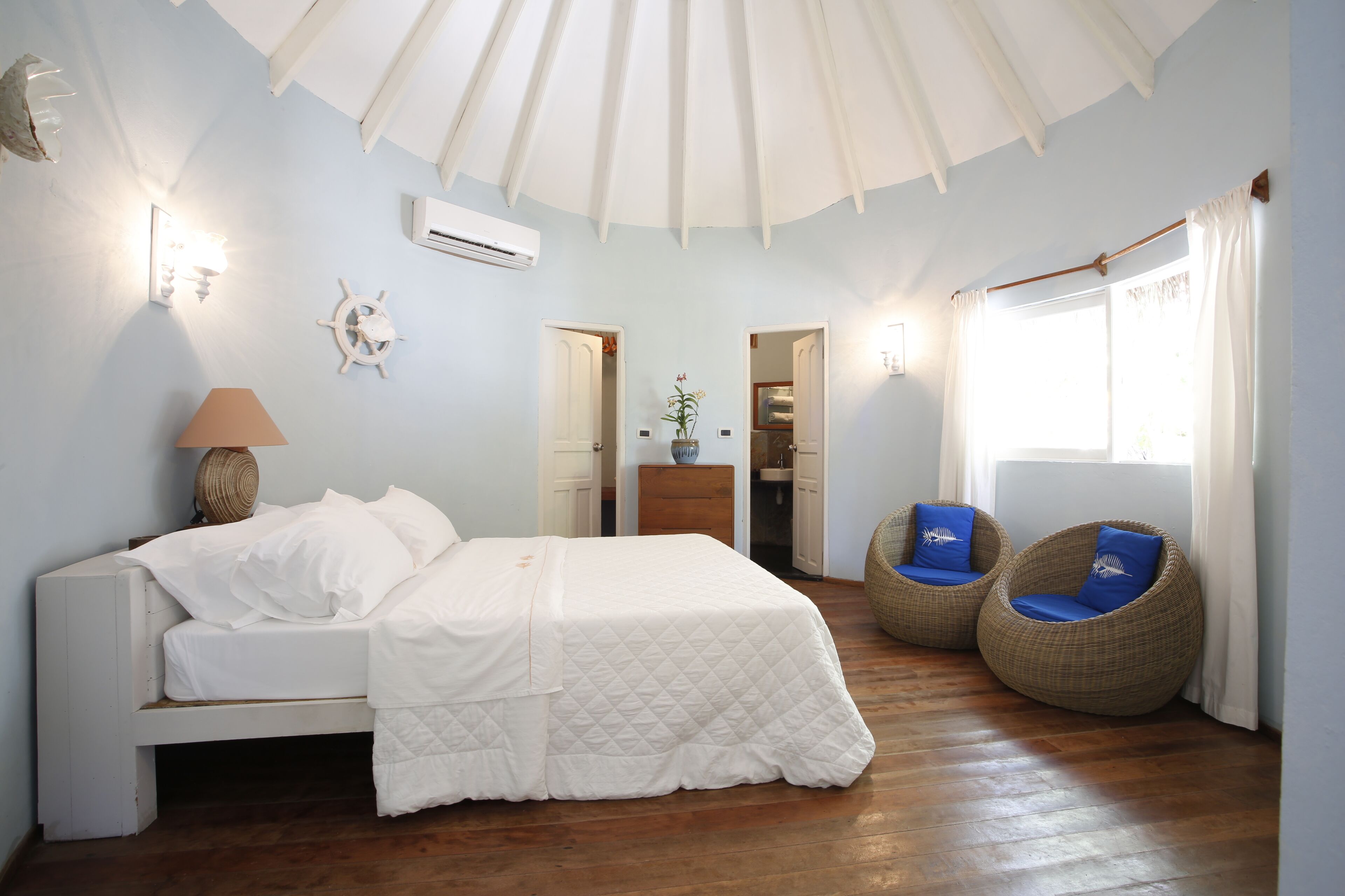 Une chambre à coucher avec un lit, deux fauteuils en osier, un ventilateur de plafond et une fenêtre avec des rideaux.