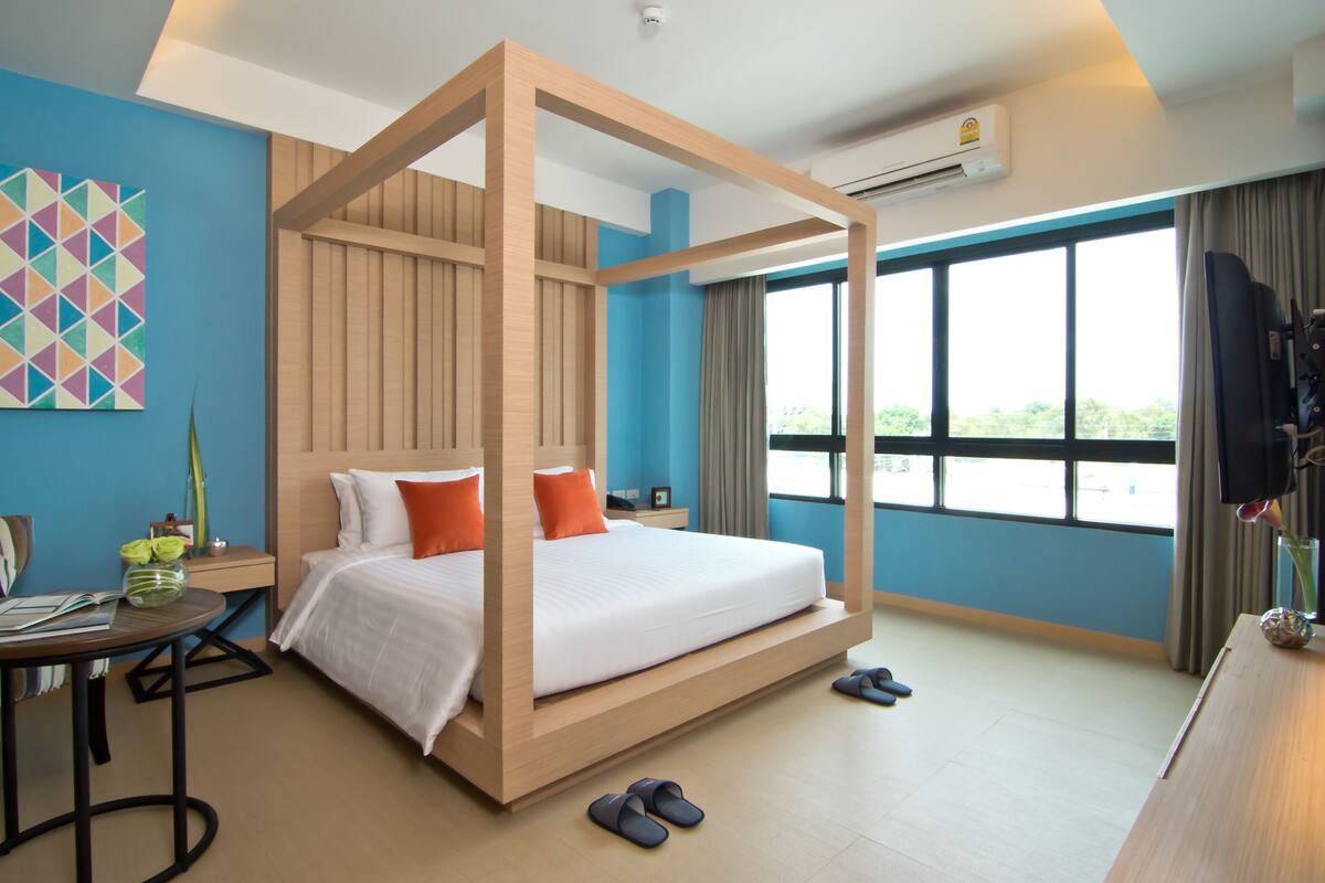 A modern hotel room with a four-poster bed, a large window with a view, a desk with a chair, and a small table with a vase of flowers.
