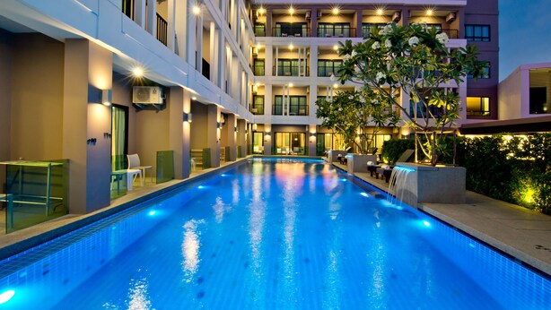 A swimming pool with a water feature, surrounded by a multi-story building with balconies and outdoor seating. - Hotel J Residence (Pattaya)
