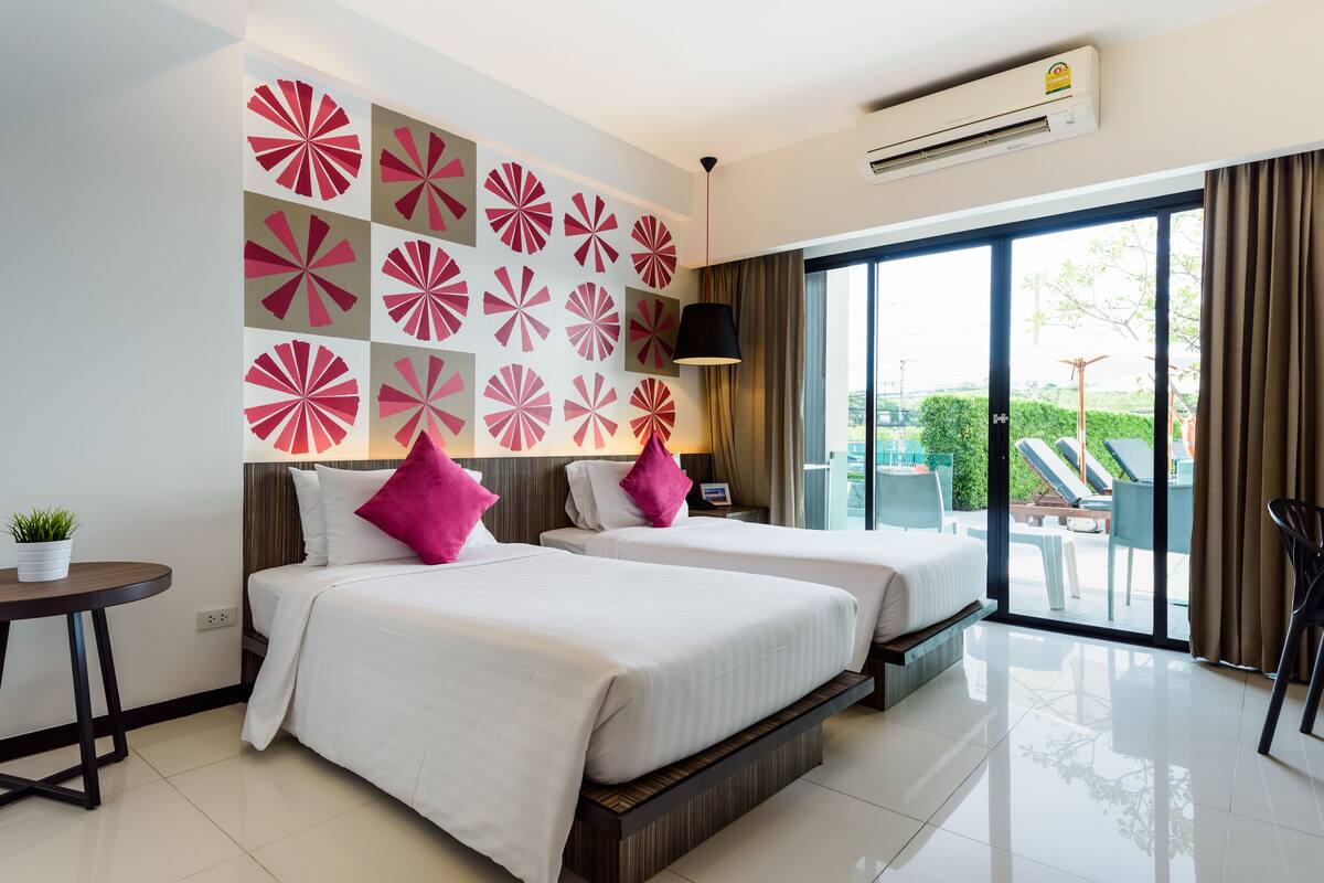 A hotel room with two beds, a small table with a plant, a wall with a geometric pattern, and a view of the outdoors through large windows.