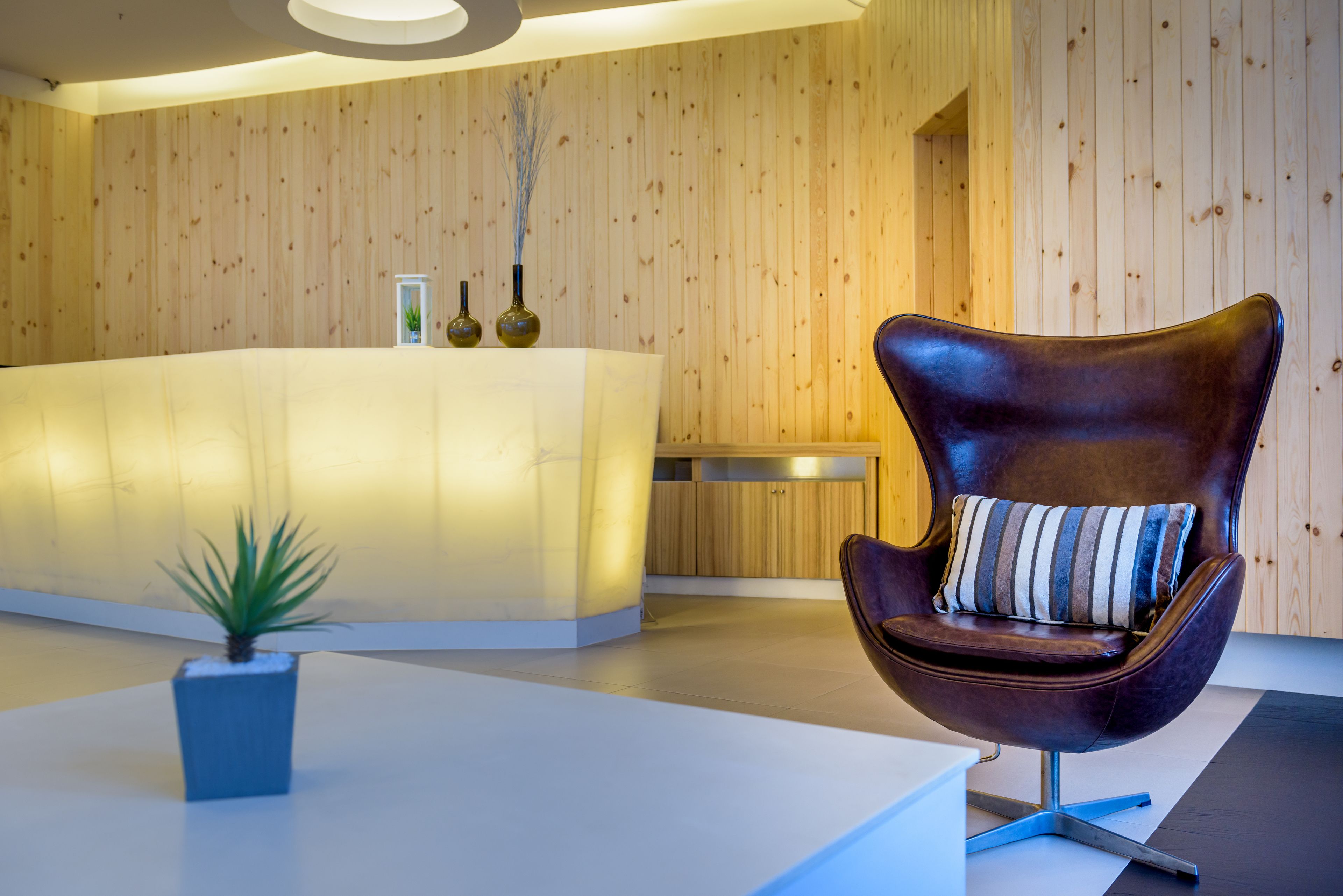 A reception area with a wooden wall, a reception desk, a modern chair with a striped pillow, and a small potted plant.