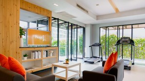 A modern hotel room with a TV, sofa, treadmill, and a view of greenery. - Hotel J Residence (Pattaya)