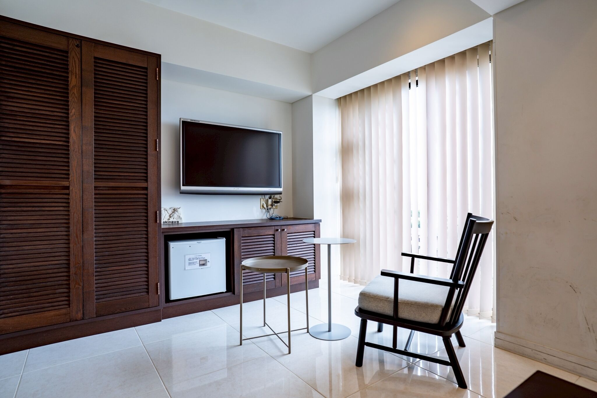 A modern living room with a flat-screen TV, a wooden cabinet, a small fridge, a round table, a chair, and a large window with curtains.