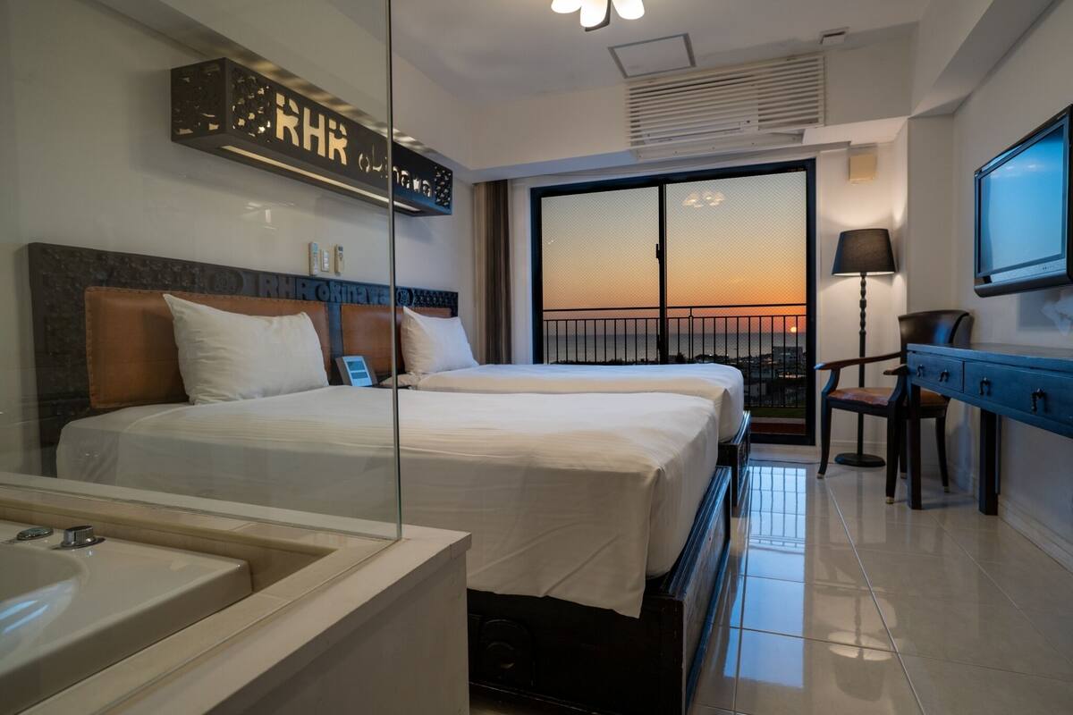 A hotel room with two beds, a large window with a view, a TV, and a desk with a lamp.