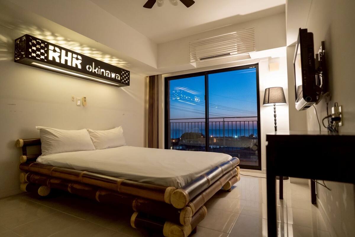 A modern hotel room with a bed, a TV, and a large window offering a view of the cityscape.