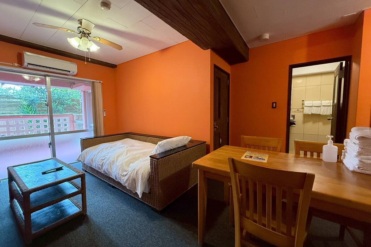 A room with a bed, a table, chairs, an air conditioning unit, a ceiling fan, and a view of the outside.