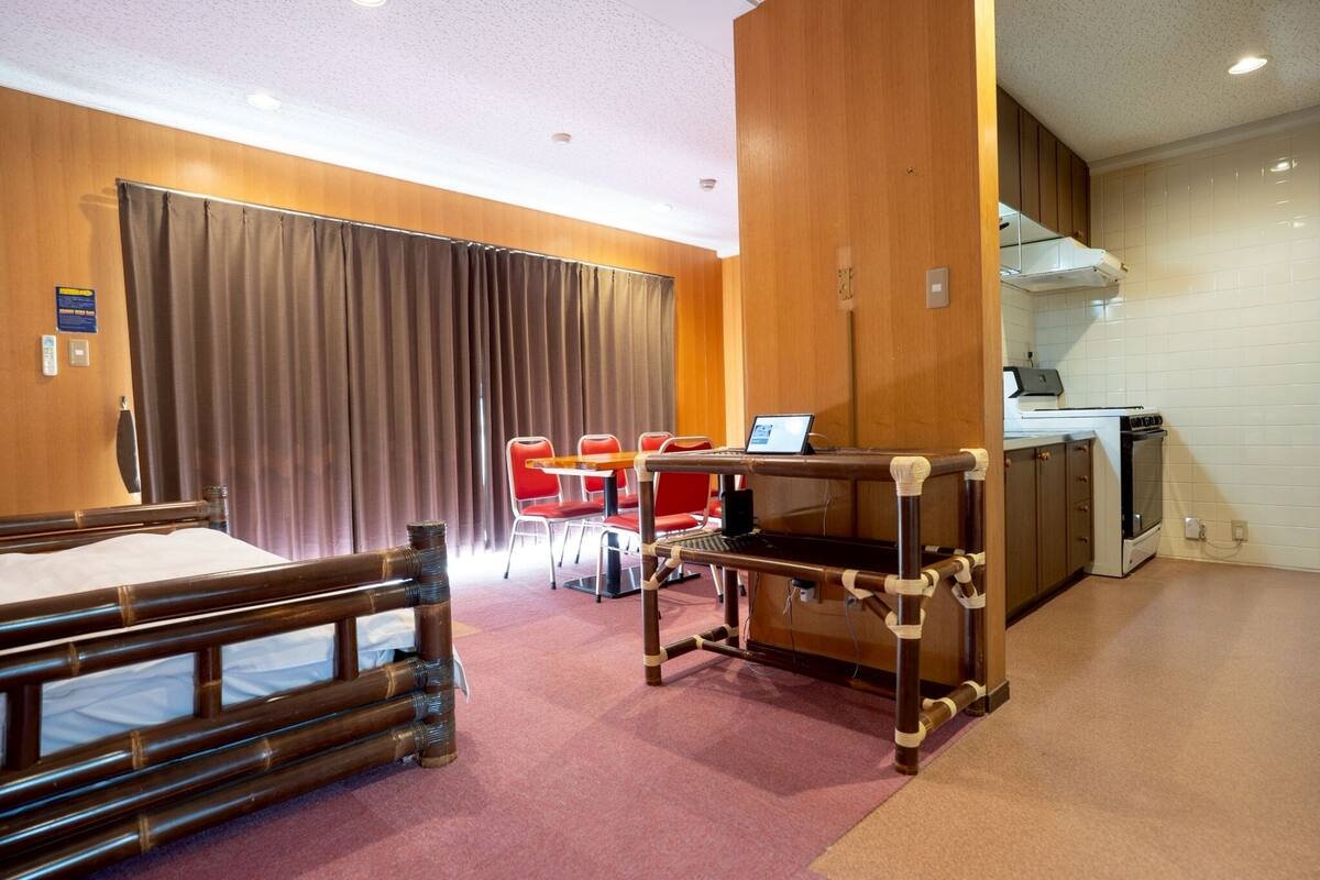 A room with a bed, a desk with a computer, red chairs, a kitchen area, and a curtain.