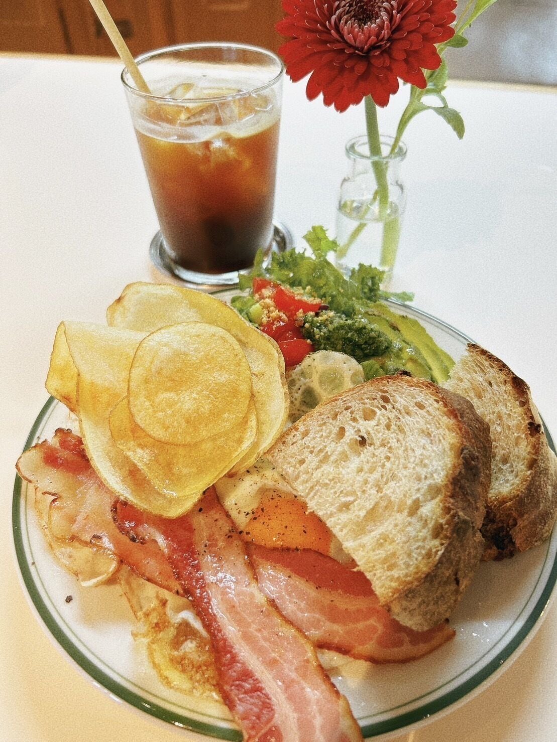 A plate with bacon, toast, chips, and salad, accompanied by a glass of iced tea and a red flower.