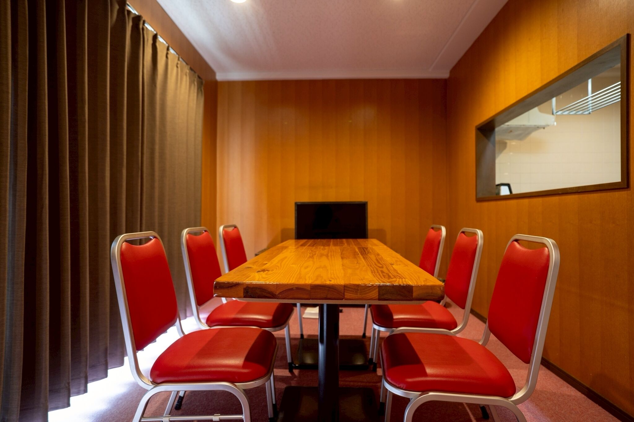 A conference room with a wooden table, red chairs, a television, and a mirror on the wall.