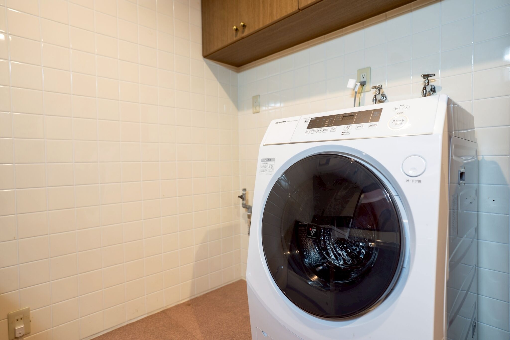 A front-loading washing machine in a laundry room with white tiled walls and a wooden cabinet above.