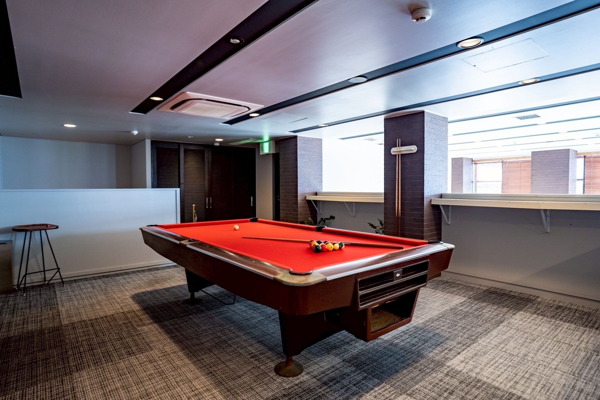 Une table de billard dans une pièce dont le sol est recouvert d’une moquette, avec un tabouret de bar et une fenêtre équipée de stores.