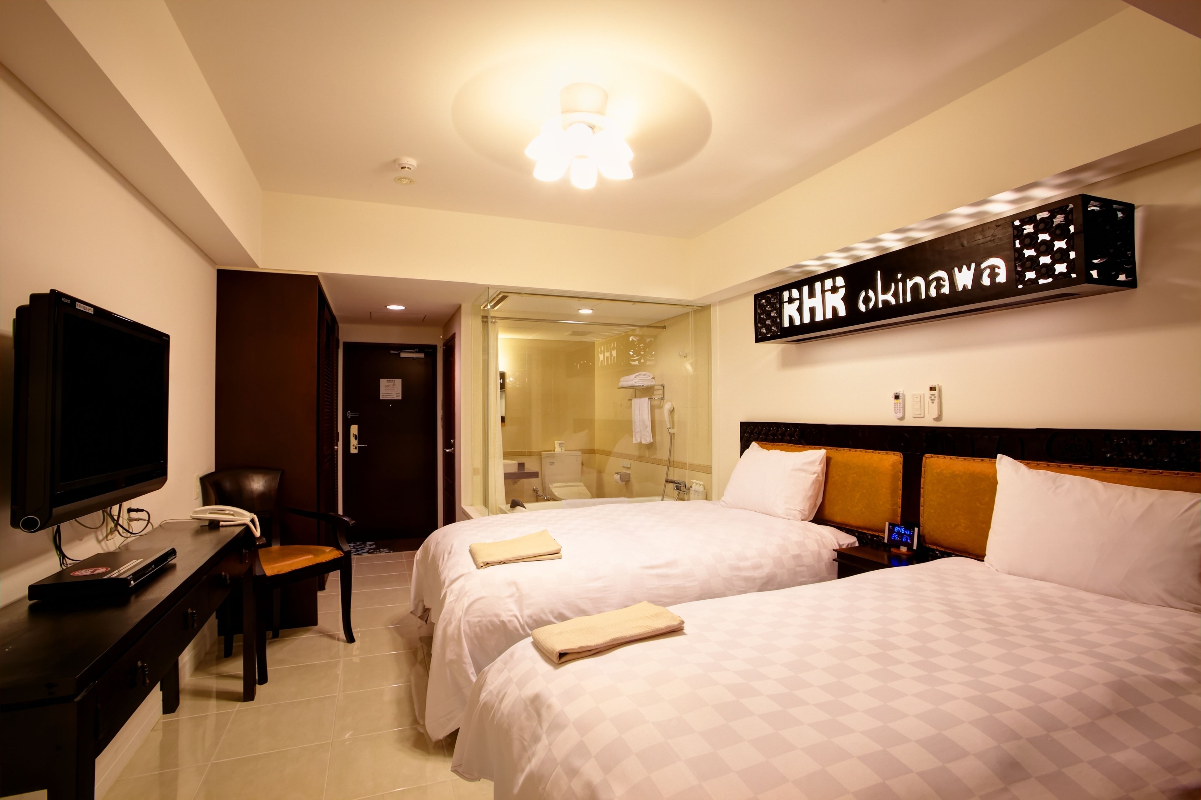 A hotel room with two beds, a TV, a desk, and a bathroom visible through a glass door.