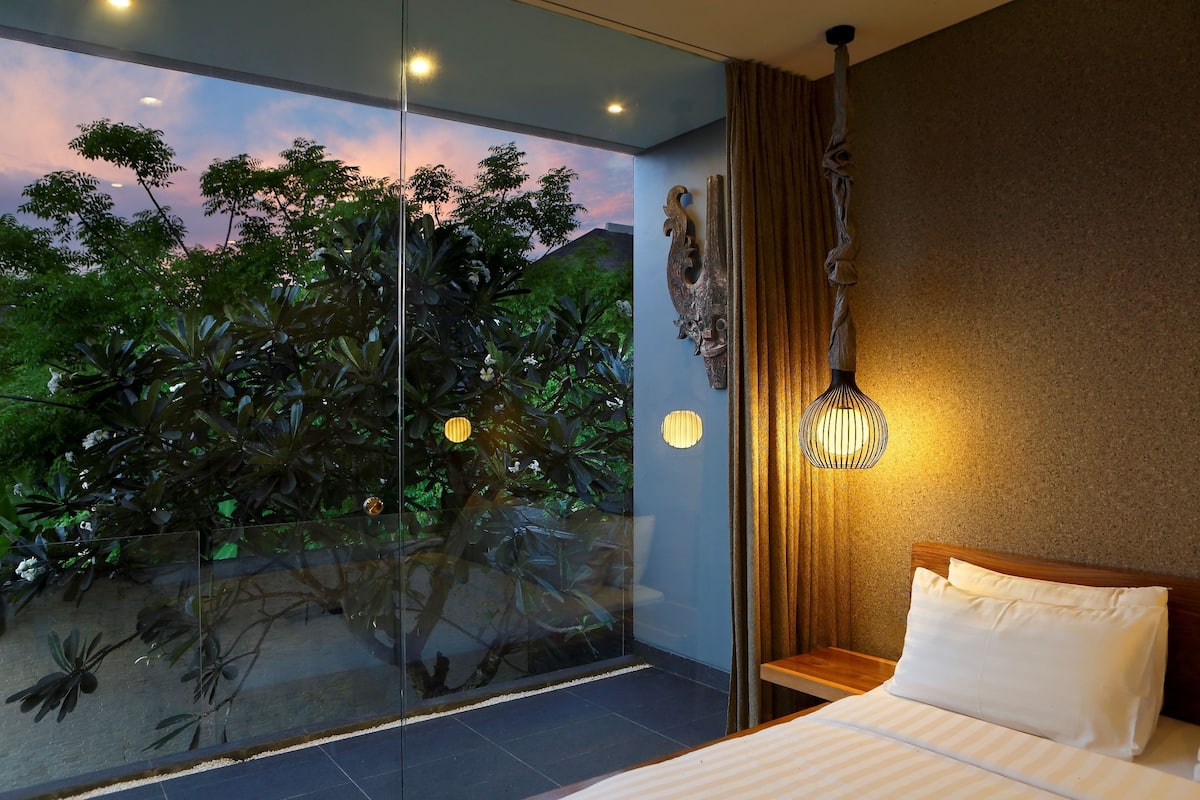 A bedroom with a large window, a bed, a nightstand, a wall-mounted light fixture, and a decorative wall piece.