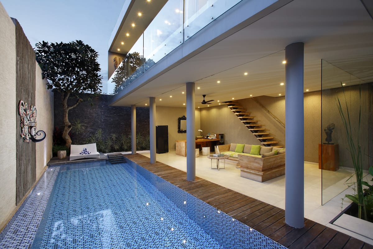 A modern outdoor pool area with a wooden deck, a seating area with a sofa and chairs, and a staircase leading to an upper level.