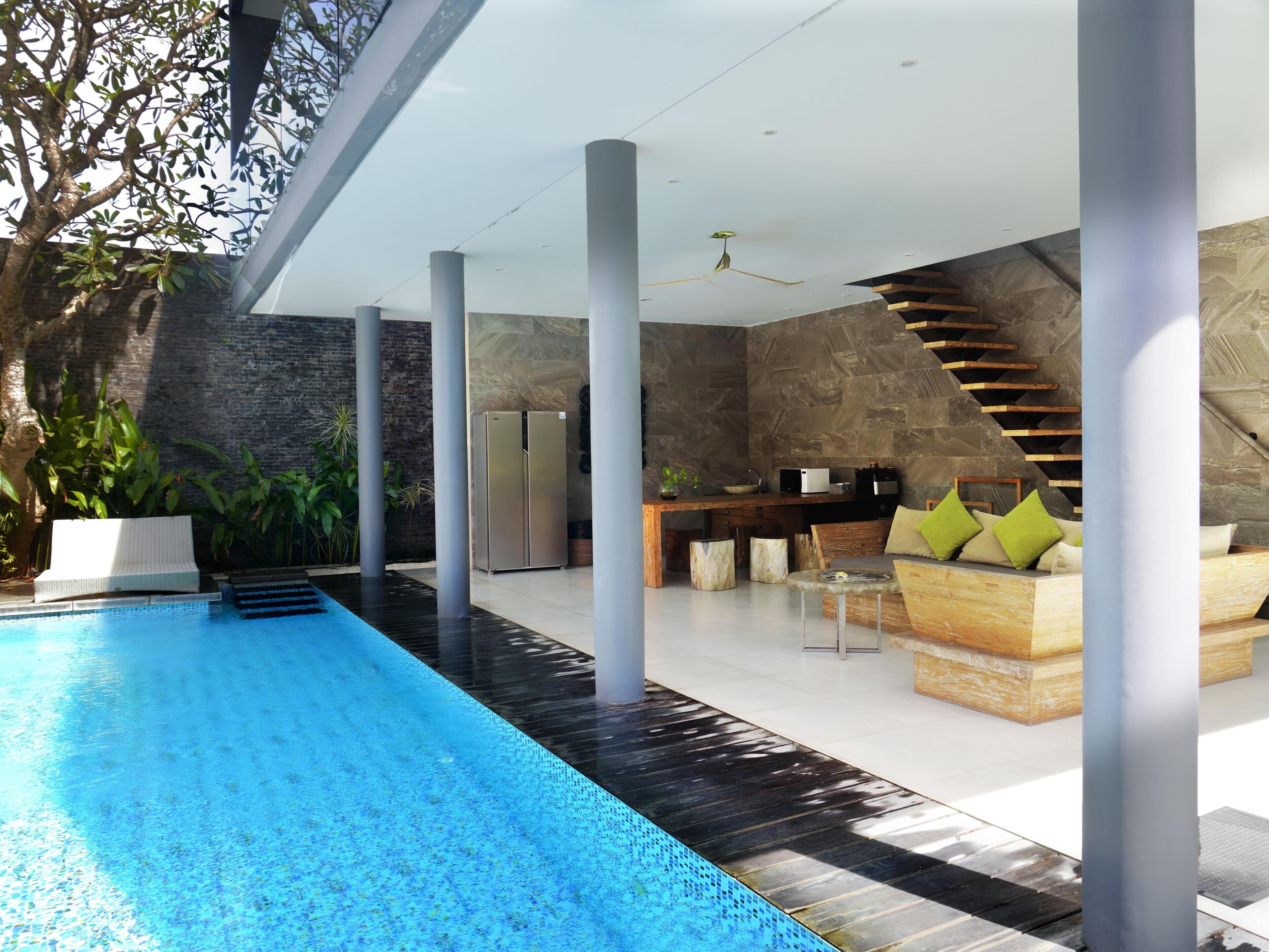Un espace piscine intérieur moderne, avec un grand bassin rectangulaire, un banc en bois agrémenté de coussins et un mur de pierre en arrière-plan.