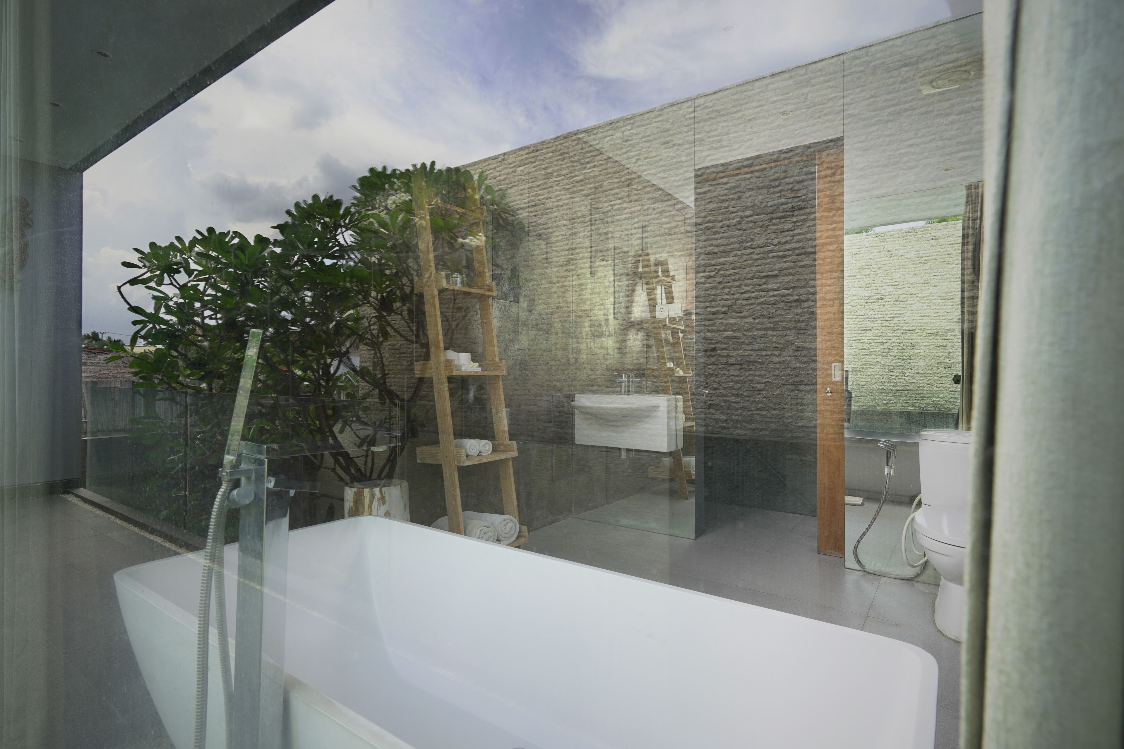 A modern bathroom with a bathtub, a wooden ladder, a sink, and a large window with a view of the sky.