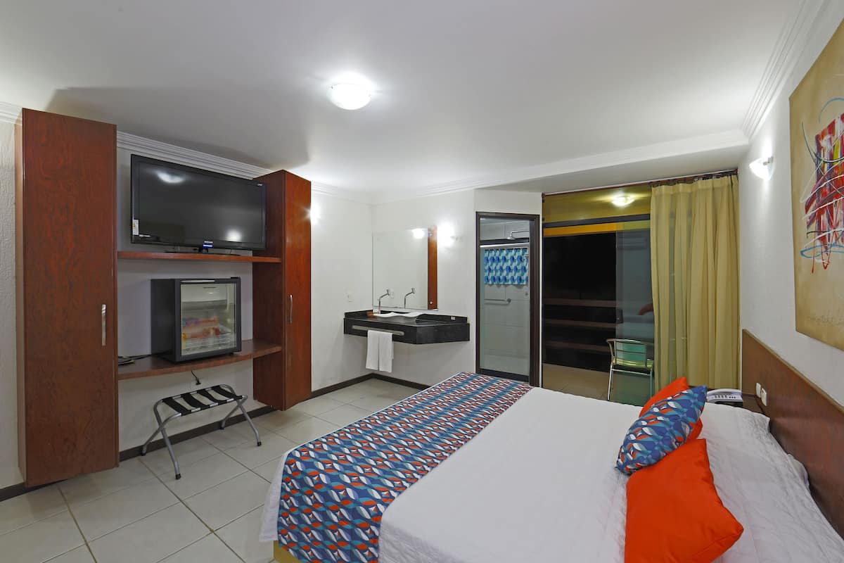 Quarto de hotel com cama, televisão, frigorífico pequeno, casa de banho e porta de correr para uma varanda.