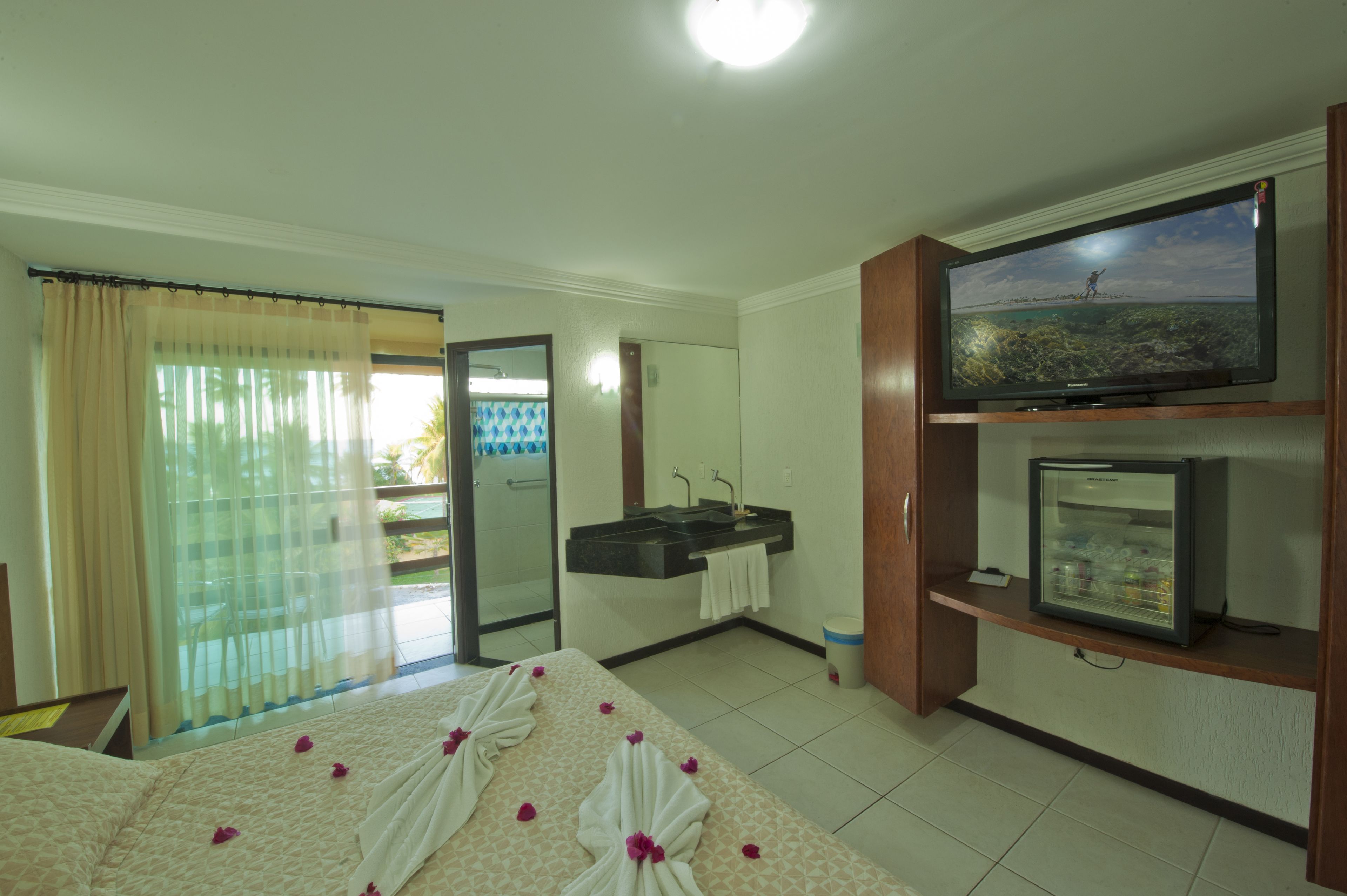 Habitación de hotel con cama, televisión, refrigerador pequeño, baño con lavamanos y espejo, y balcón con cortinas.