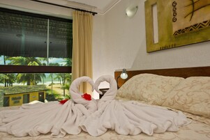 Habitación de hotel con cama, ventana que da a palmeras y una playa, y un cuadro colgado en la pared.