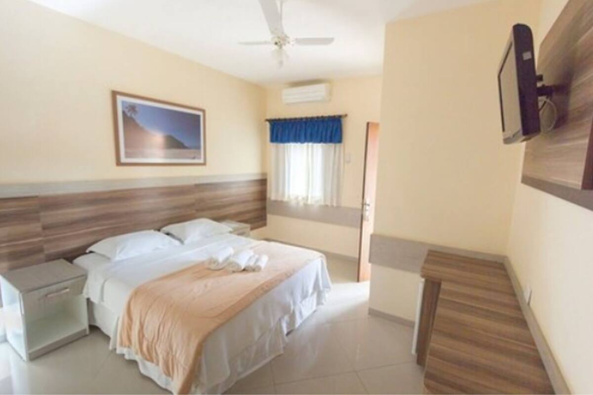 A hotel room with a bed, a nightstand, a ceiling fan, a mounted TV, and a window with curtains.