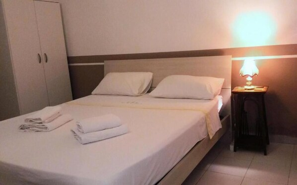 Una cama bien hecha con sábanas y almohadas blancas, una lámpara de noche y un armario en una habitación de hotel.
