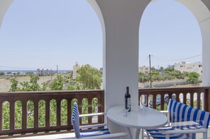 A balcony with a white table and blue-striped sofa, overlooking a coastal landscape with buildings and greenery. - Margarita Studios (Santorini)