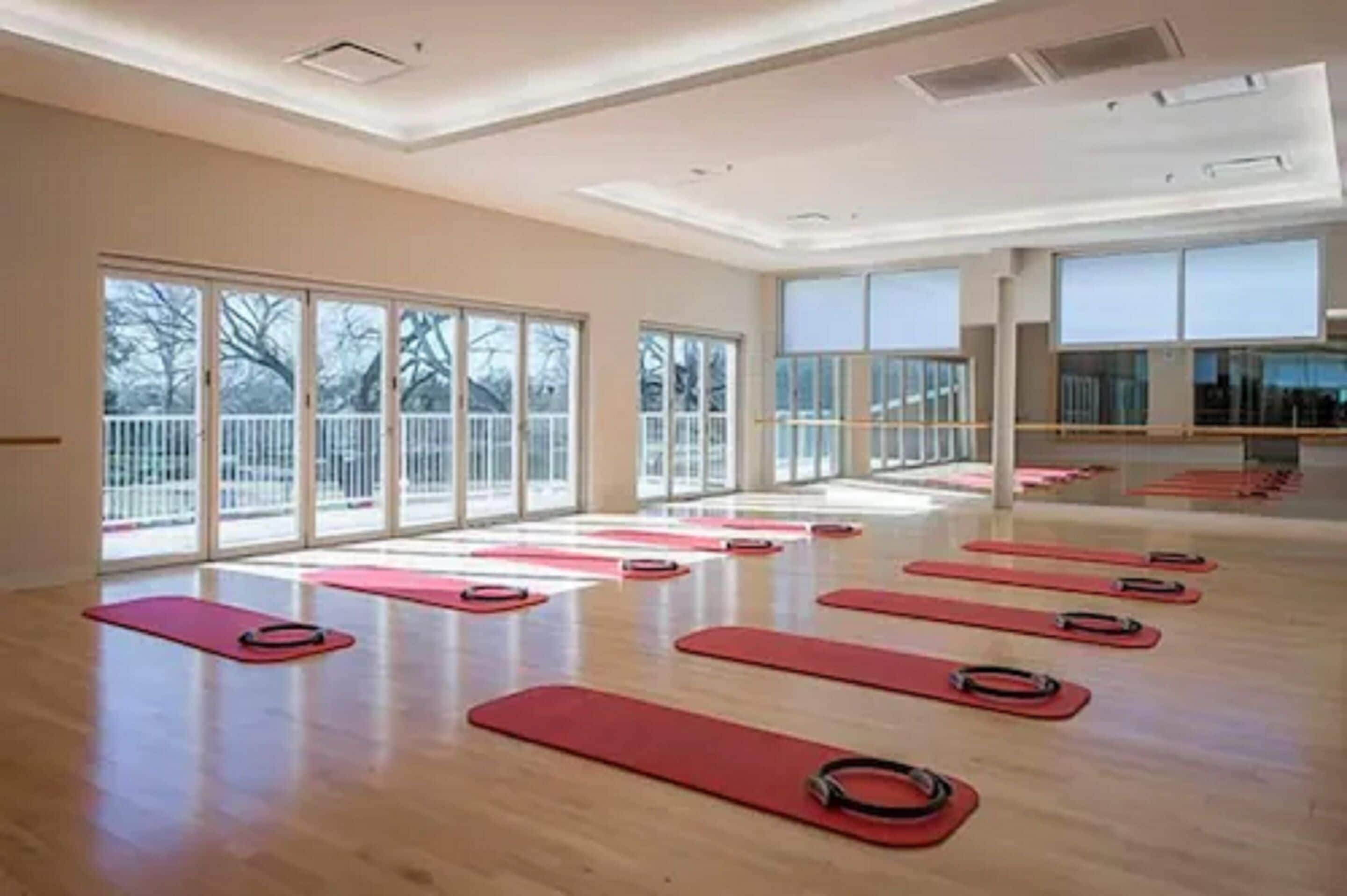 A spacious room with large windows, wooden floors, and red exercise mats arranged for group activities.