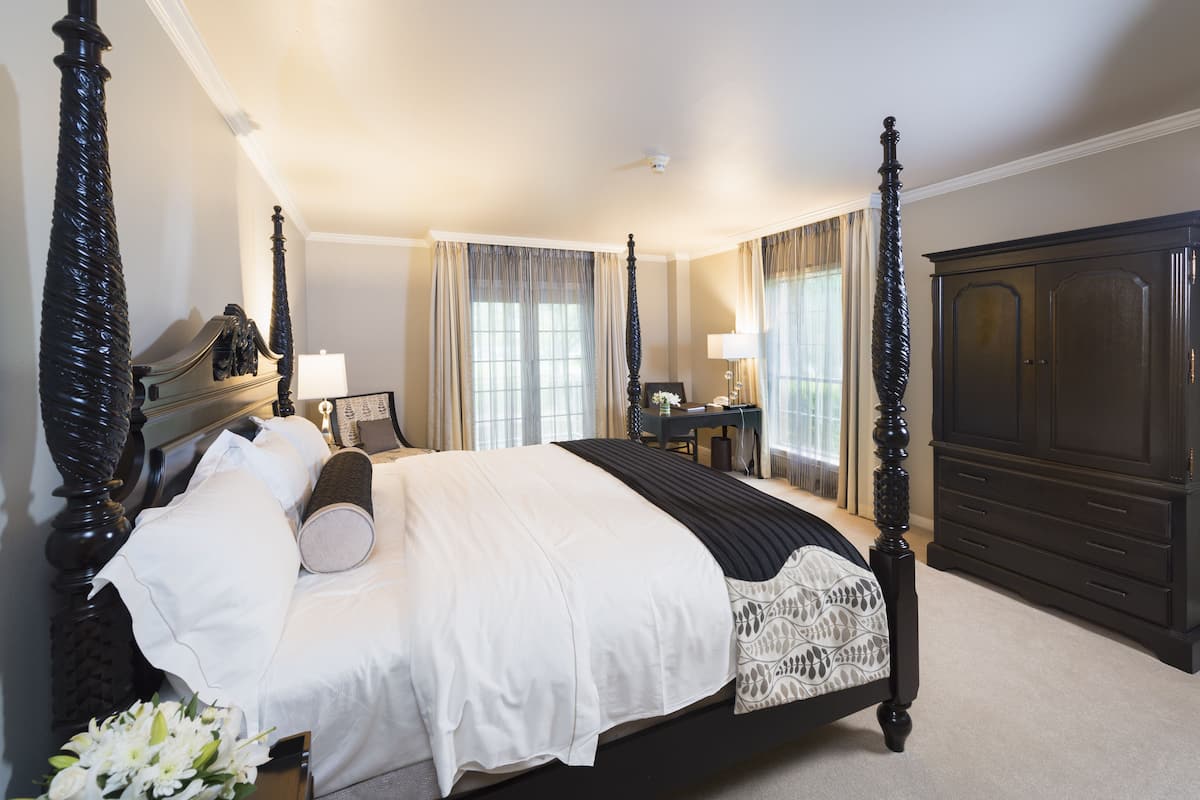 A four-poster bed with a white and black bedspread, a nightstand with a lamp, and a dark wooden wardrobe.