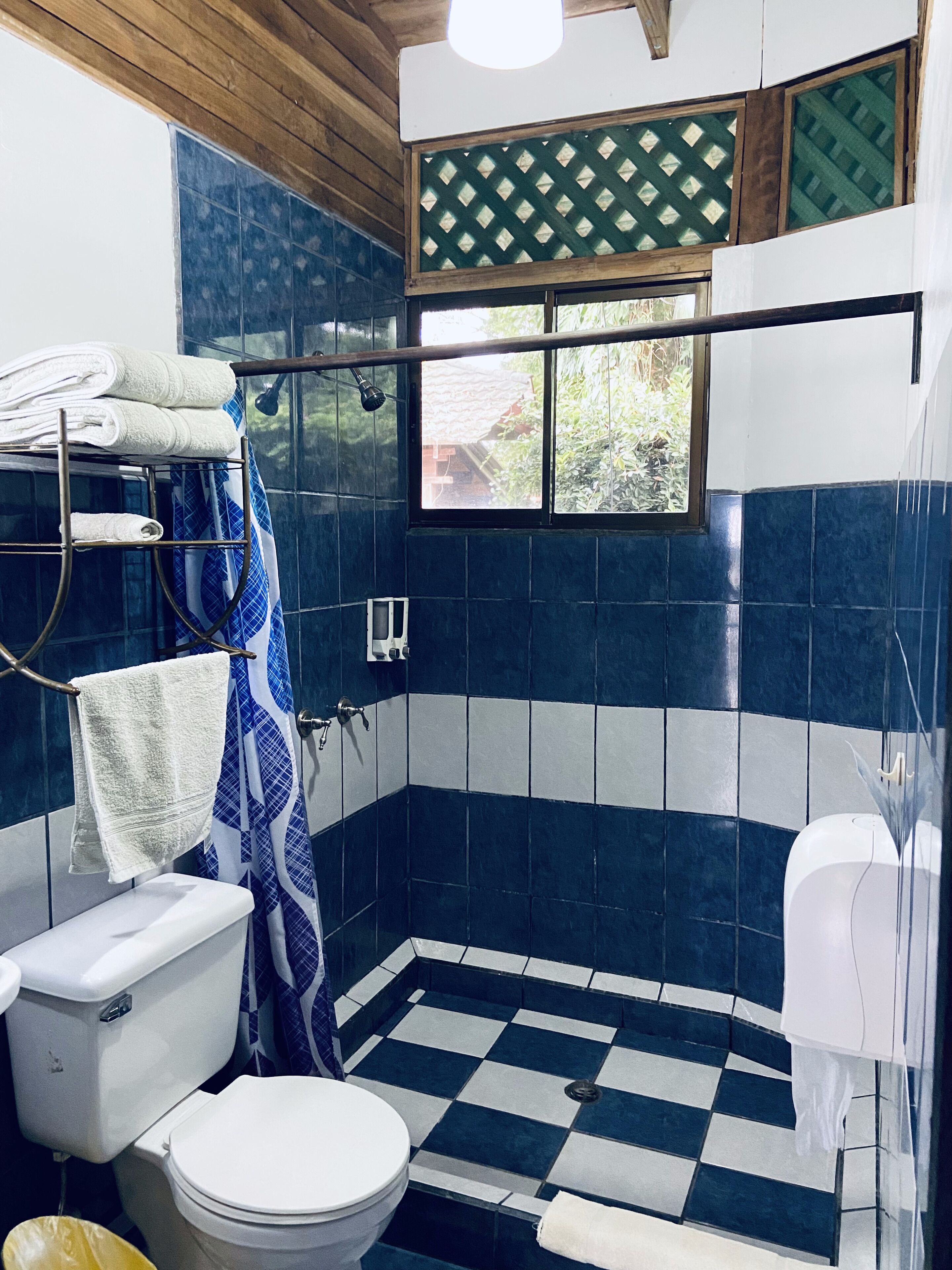 Ein Badezimmer mit Schachbrettboden, einer Toilette, einer Duschkabine mit Vorhang und Handtüchern auf einem Ablagekorb.