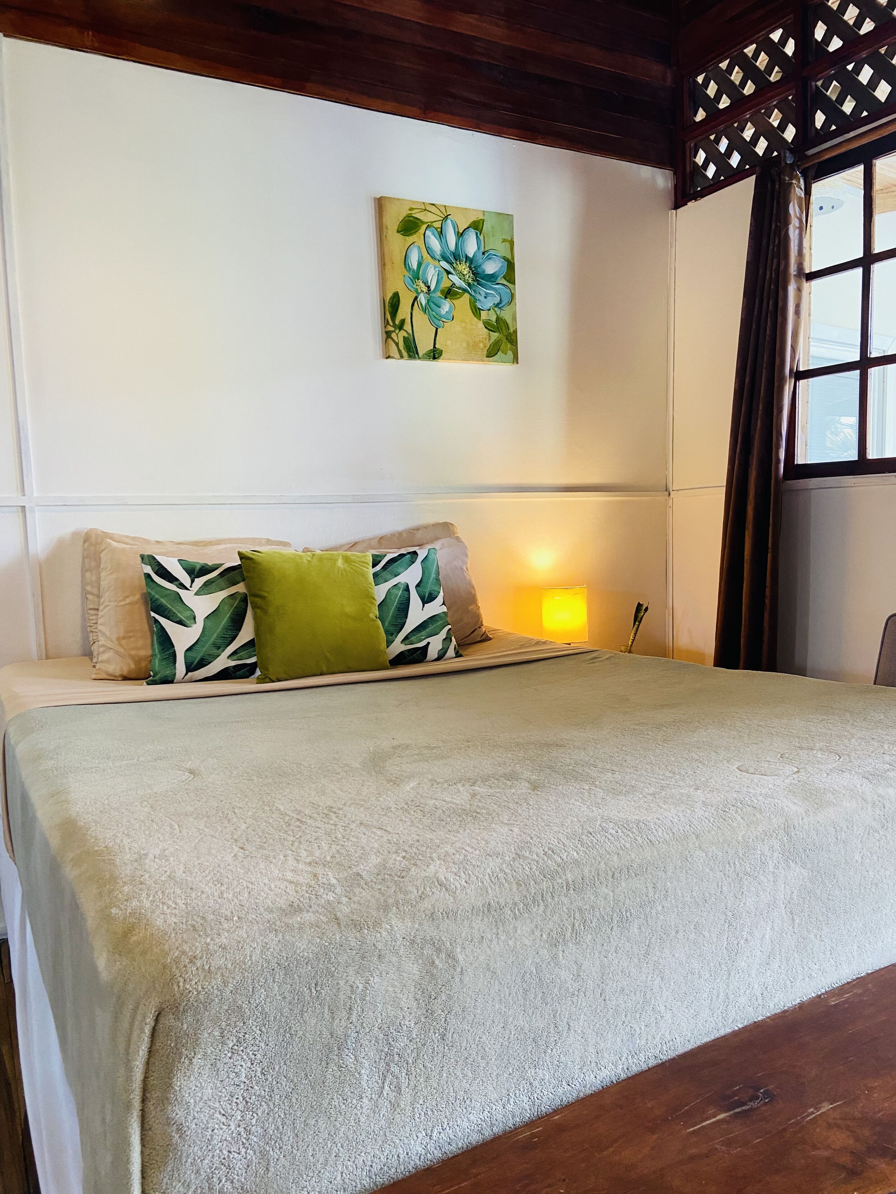 Een slaapkamer met een bed, kussens, een schilderij en een raam met gordijnen.