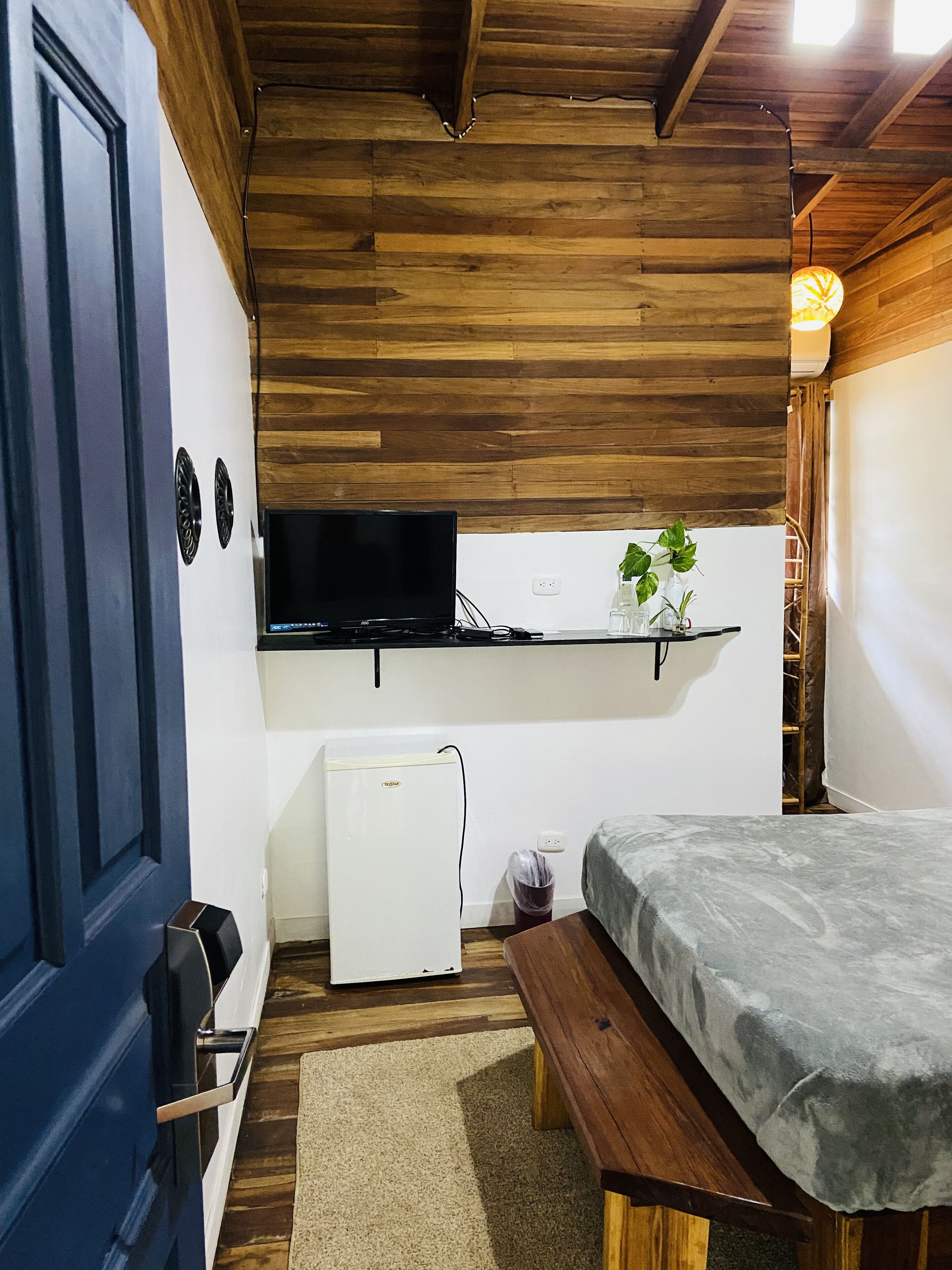 A compact room with a bed, a small white refrigerator, a TV mounted on the wall, and a wooden bench.