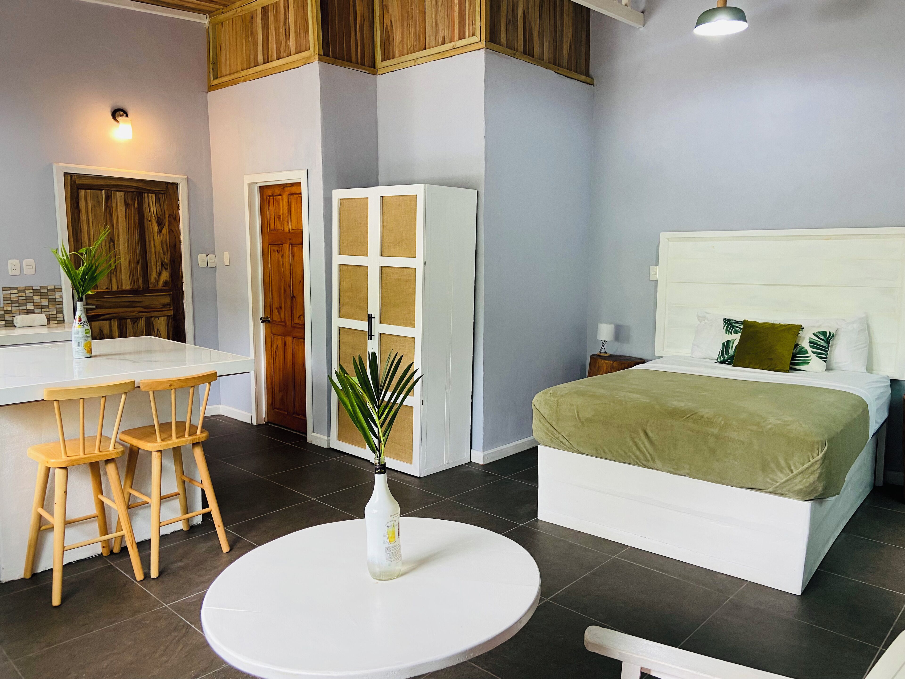 Un dormitorio con cama, una mesita redonda con una botella, dos sillas y una planta.