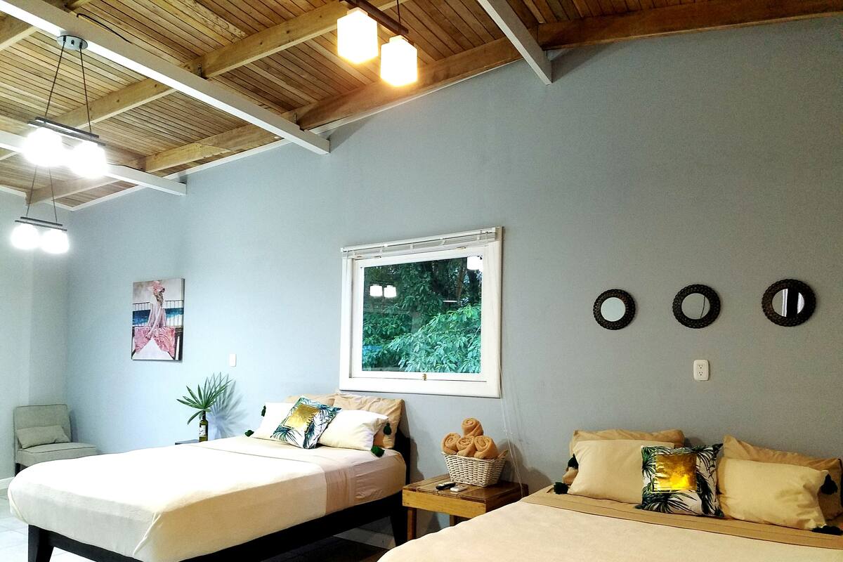 A room with two beds, a window with a view of greenery, a painting, and a ceiling with wooden beams and lights.