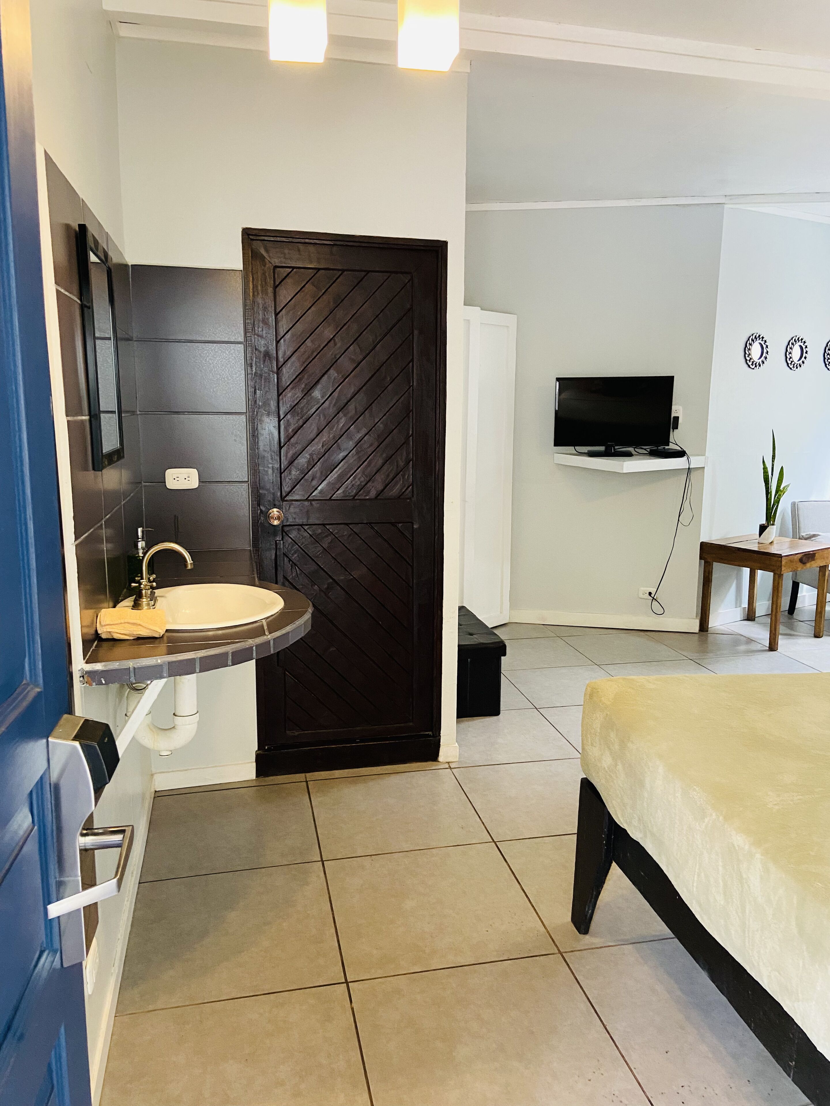 Ein Hotelzimmer mit einer dunkelbraunen Holztür, einem weißen Waschbecken, einem Bett mit einer beige farbenen Bettdecke, einem Fernseher auf einem weißen Regal und einem kleinen Tisch mit einer Pflanze.