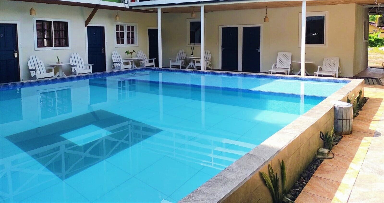 A clear blue swimming pool with white lounge chairs and tables, surrounded by a covered patio area with doors and windows.