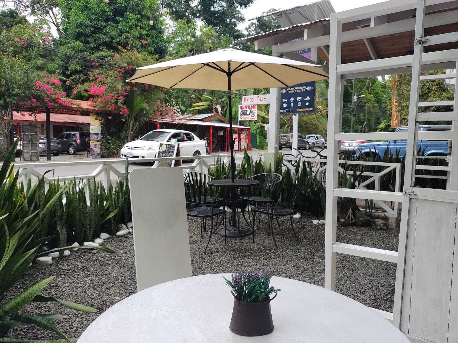 Area esterna con tavoli e sedie, un ombrellone grande e un cartello per la colazione.