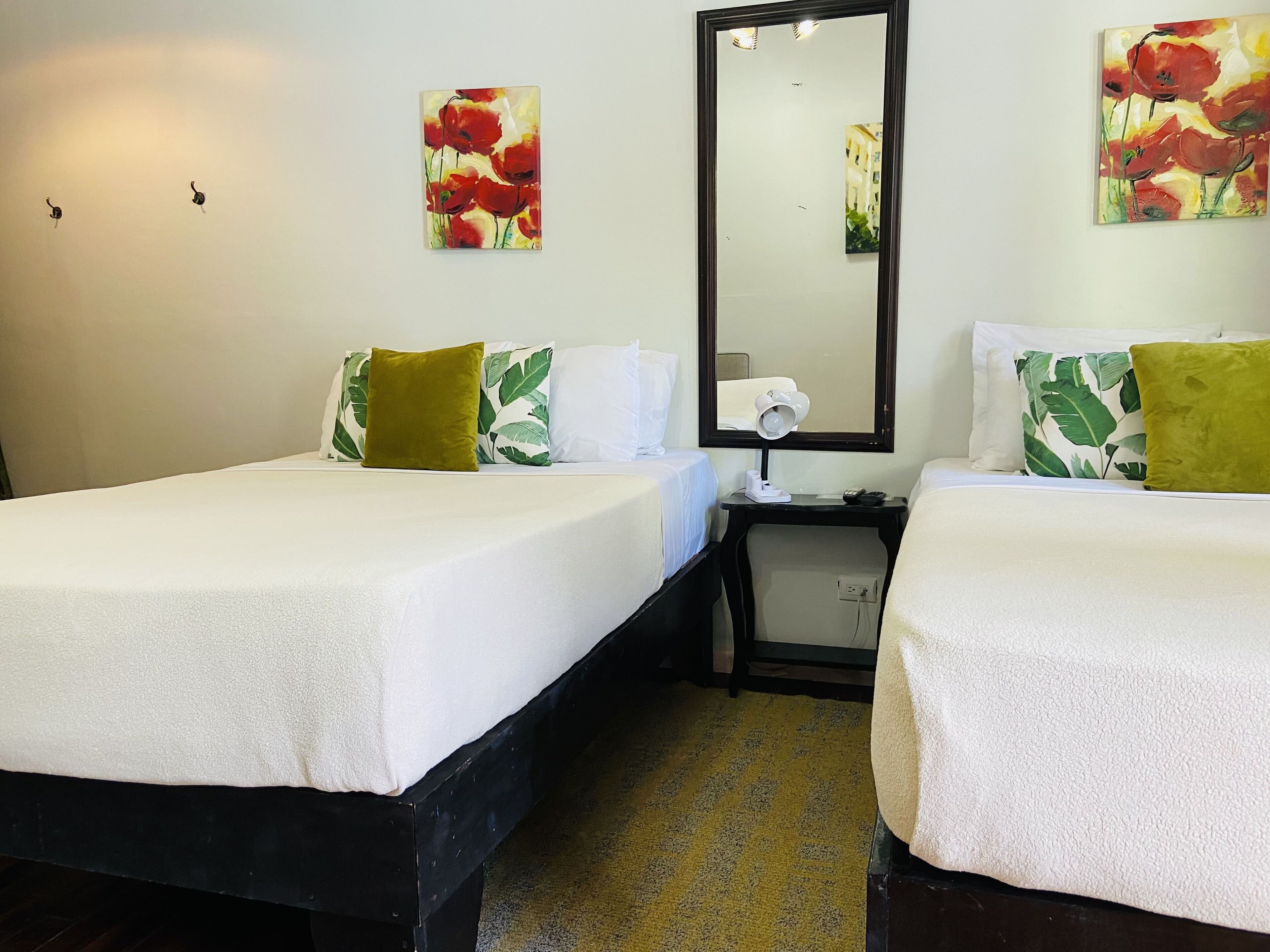 A hotel room with two beds, each with white bedding and green pillows, a nightstand with a lamp, and a mirror on the wall.