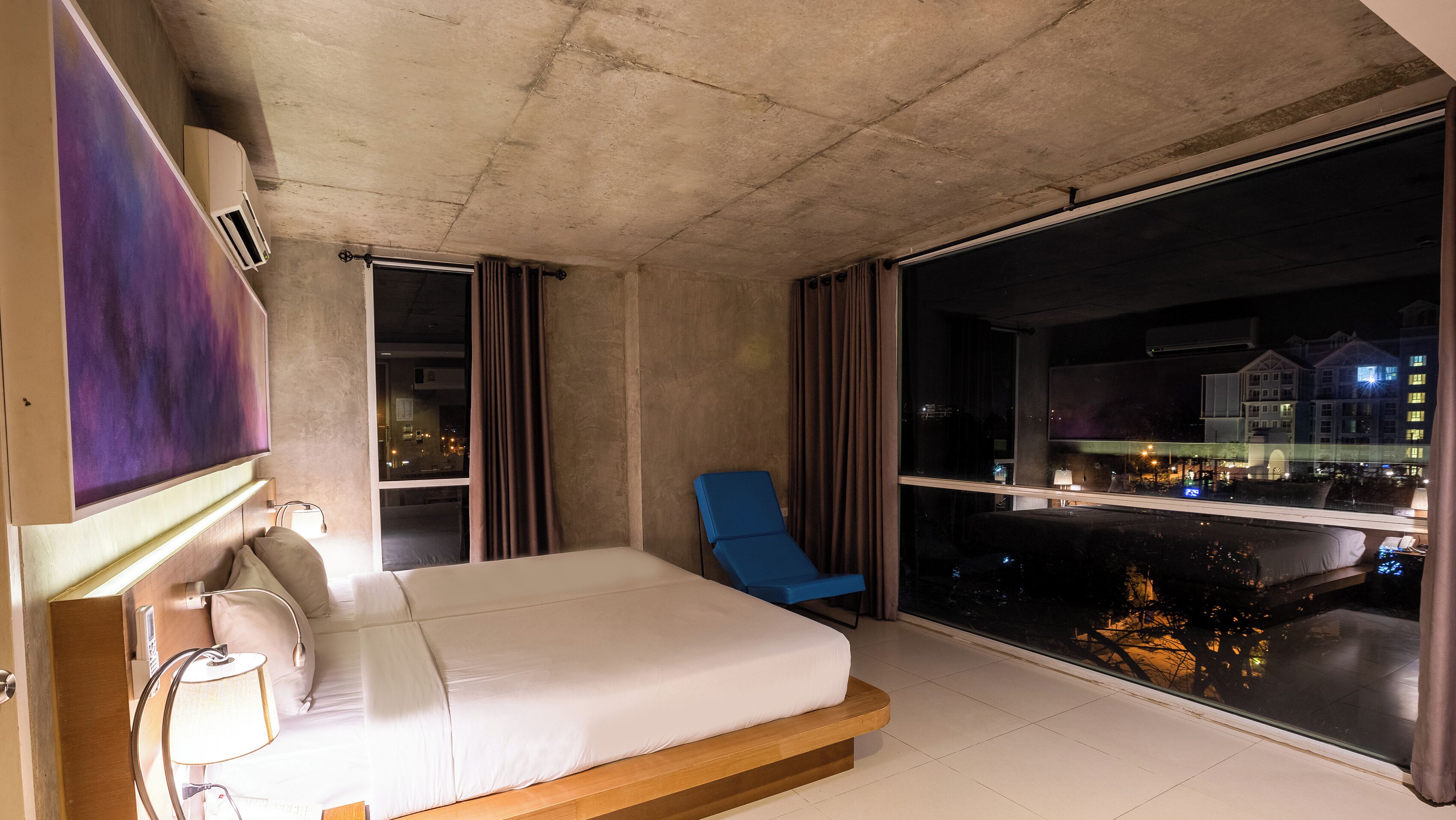 A hotel room with a bed, a chair, a large window with a view of the city, and a mounted TV.