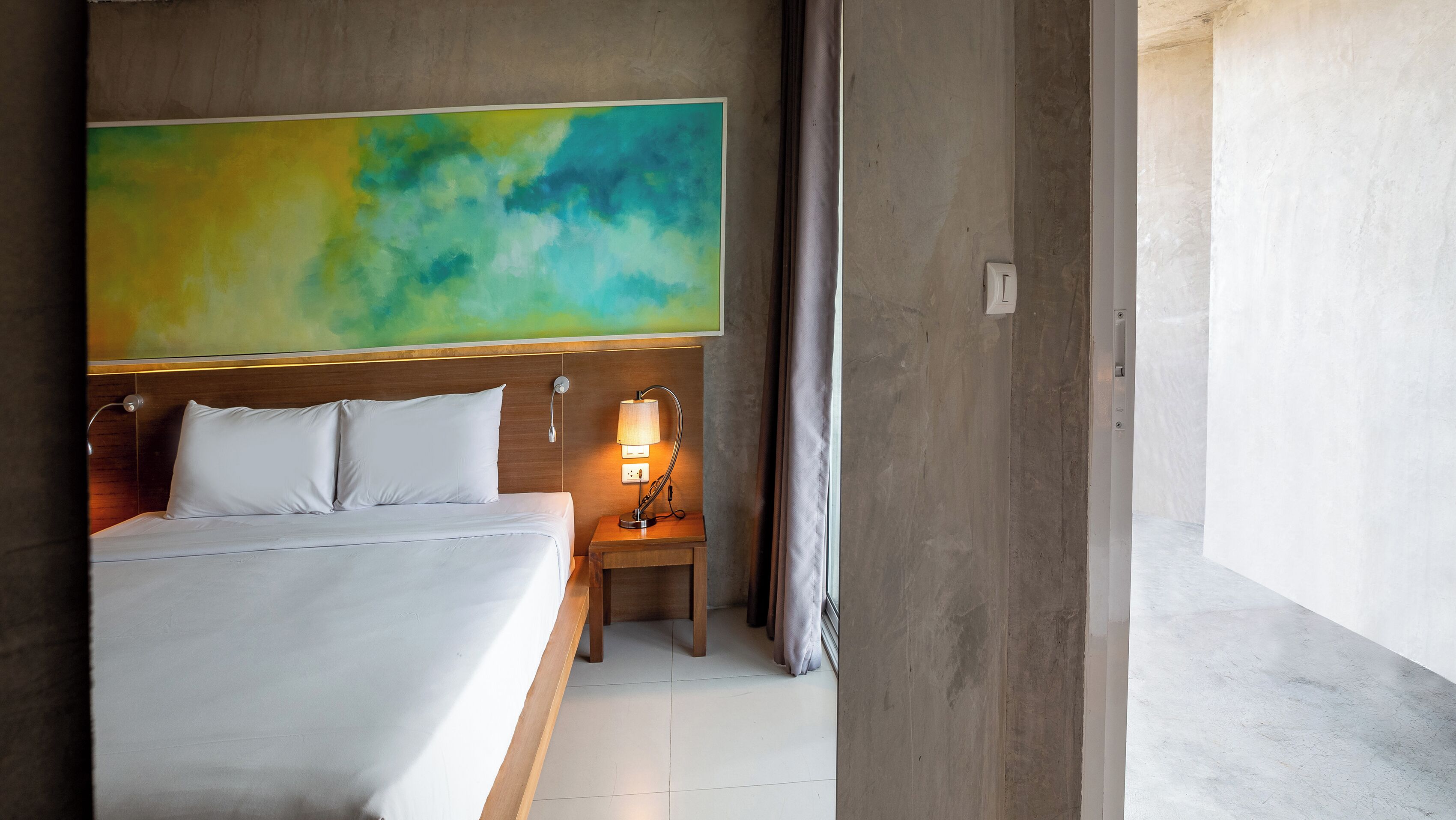 Een hotelkamer met een groot bed, een nachtkastje met een lamp en een groot abstract schilderij aan de muur.