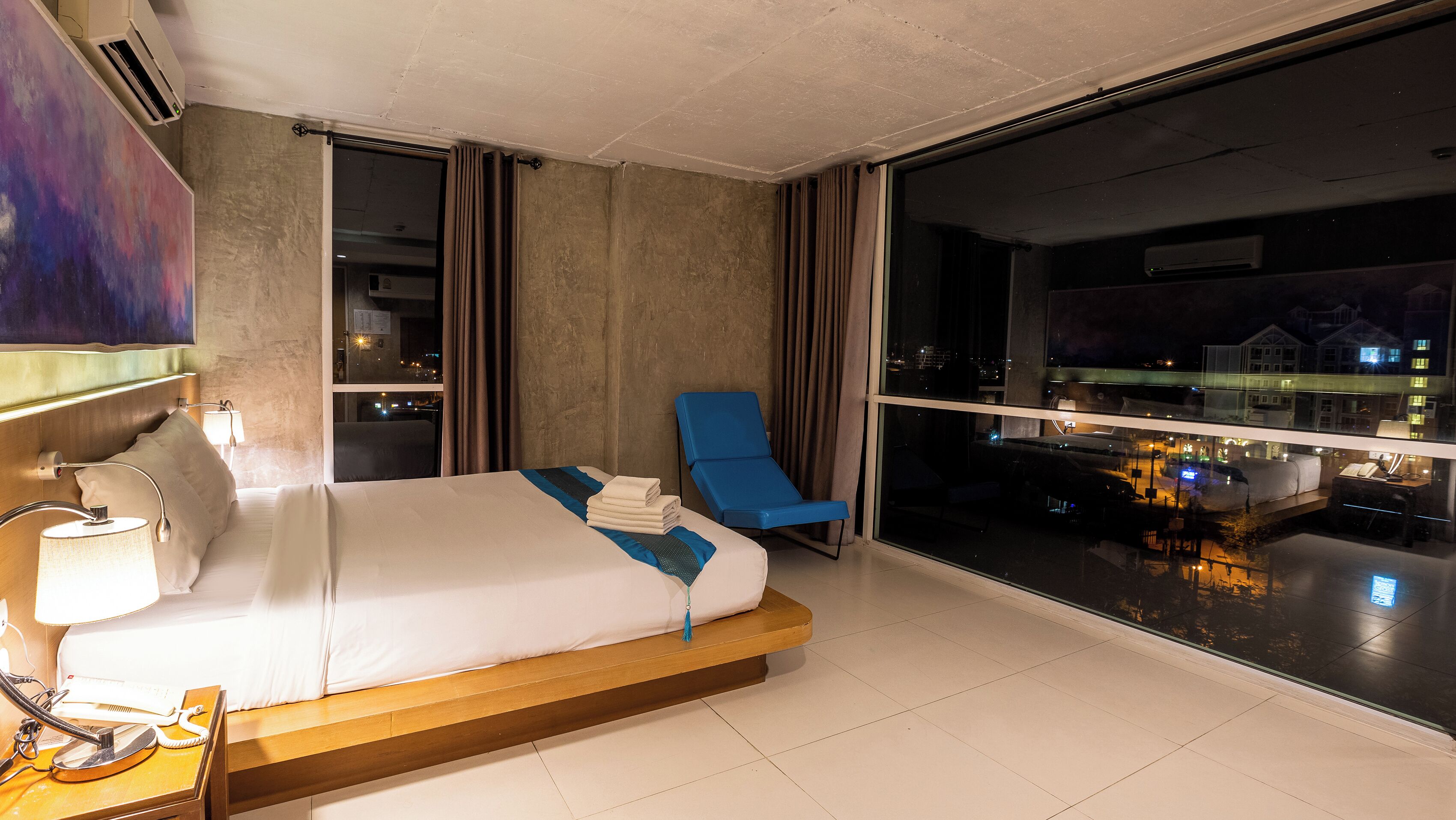 Habitación de hotel con una cama, una silla azul, una ventana con vista a la ciudad y una mesita de noche con lámpara y teléfono.
