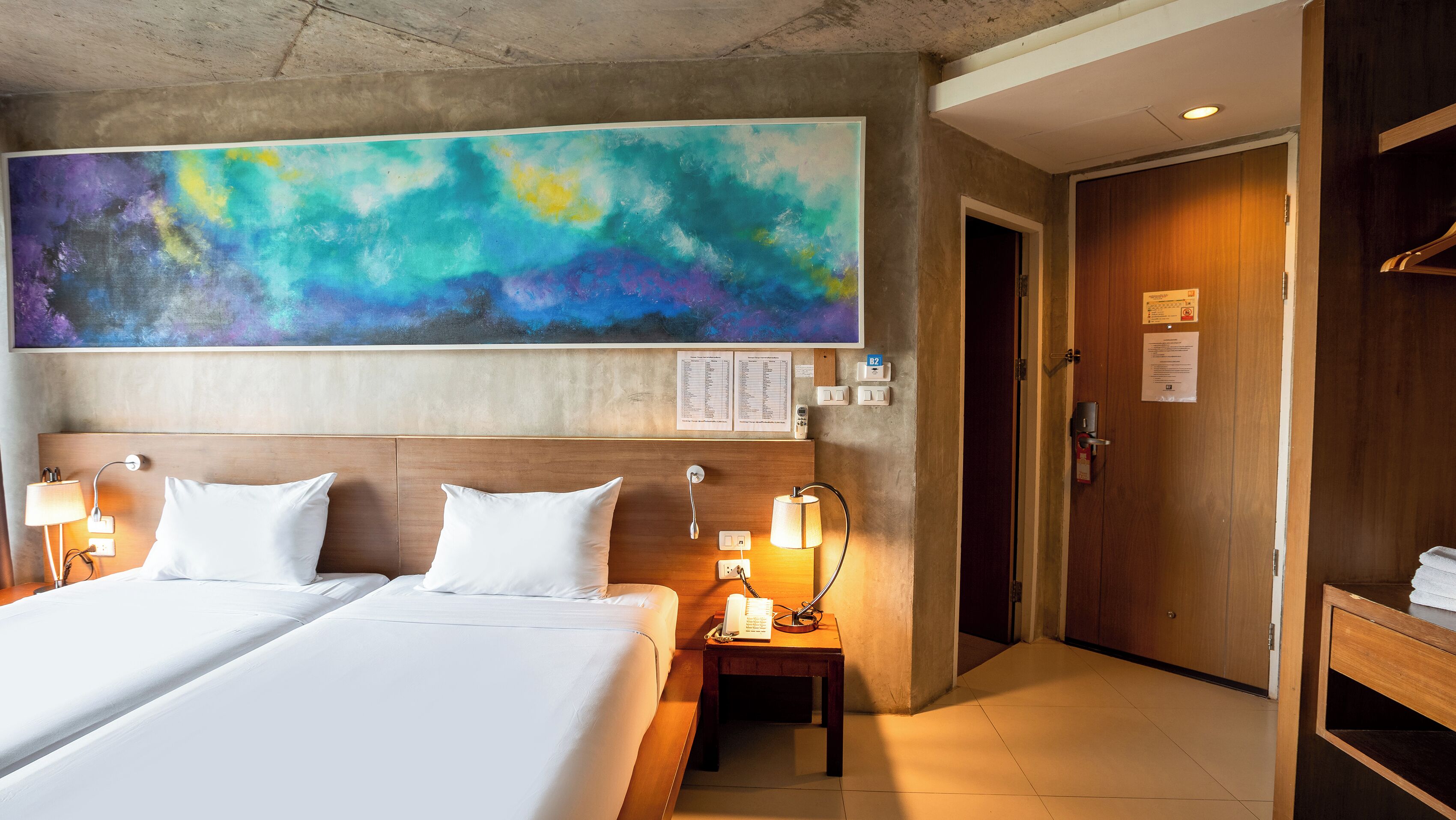 Habitación de hotel con un cuadro abstracto grande, una cama doble con ropa blanca, dos mesitas de noche con lámparas y una puerta de madera.