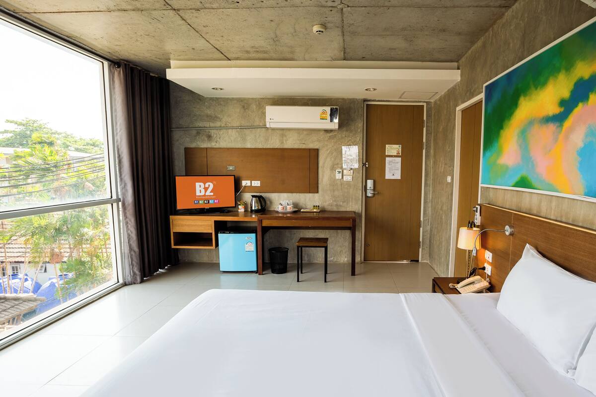 A hotel room with a bed, a desk with a computer, a small fridge, a window with curtains, and a colorful painting on the wall.