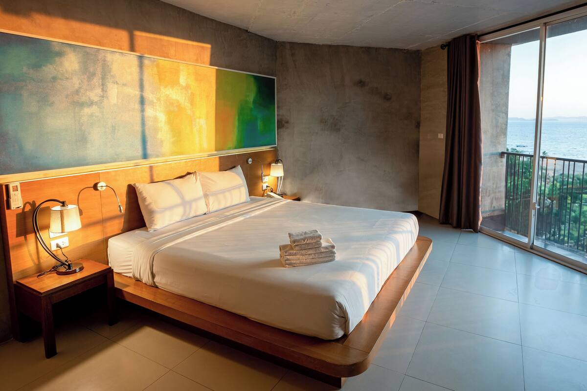 A modern hotel room with a large bed, bedside lamps, a painting on the wall, and a balcony with a view of the sea.