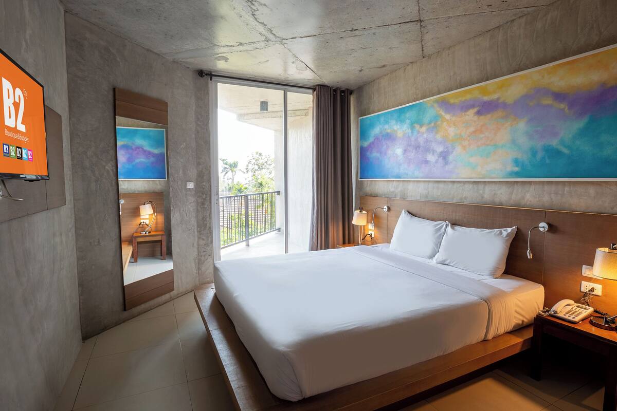 A hotel room with a large bed, a painting above the bed, a desk with a lamp, a phone, and a TV displaying 'B2 Boutique & Budget'.