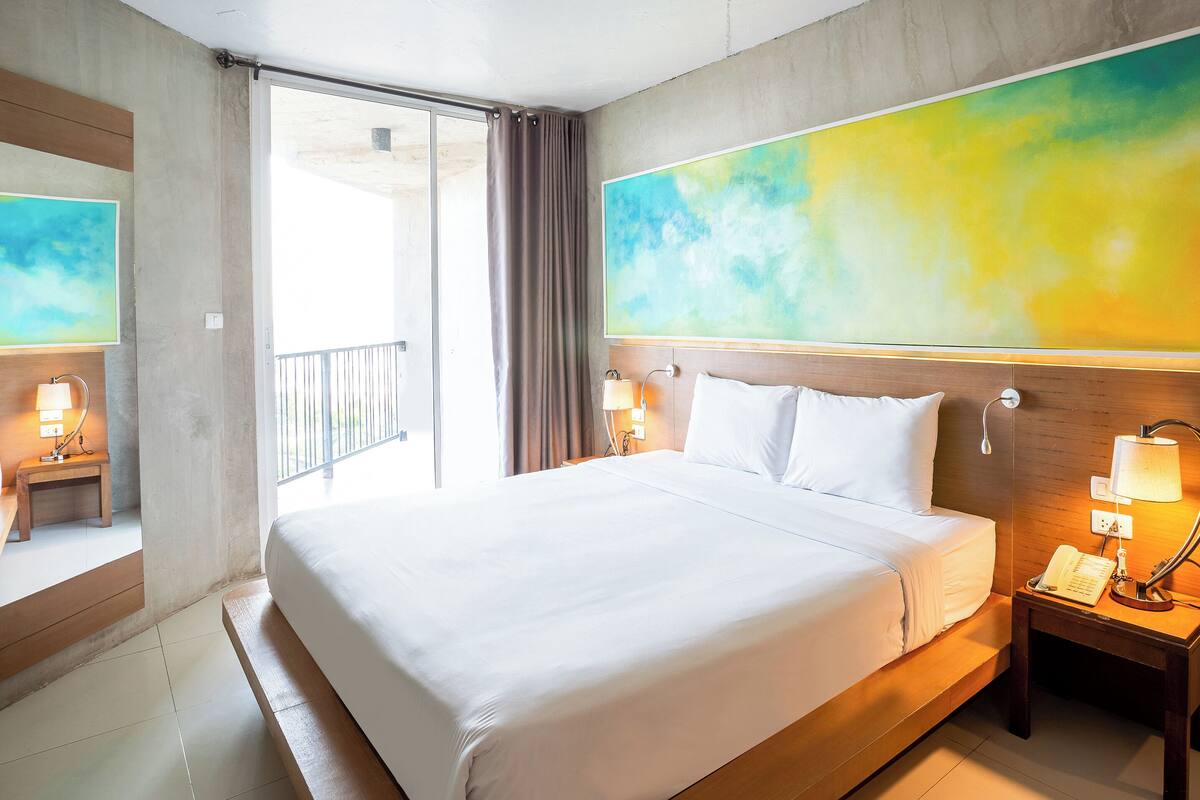 A hotel room with a large bed, a nightstand with a lamp, a mirror, and a large abstract painting on the wall.