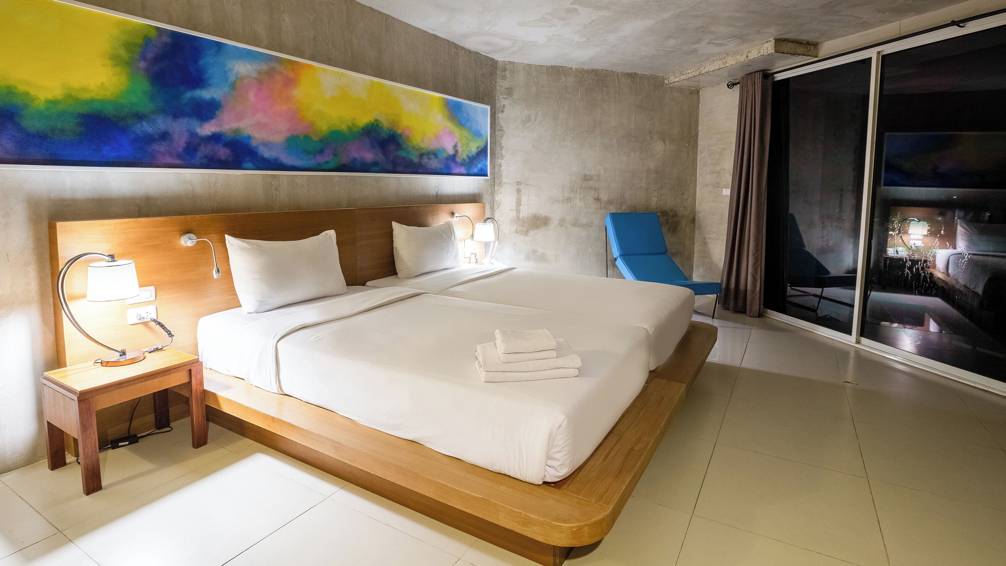 A modern hotel room with a large bed, bedside lamps, a blue chair, and a large abstract painting on the wall.