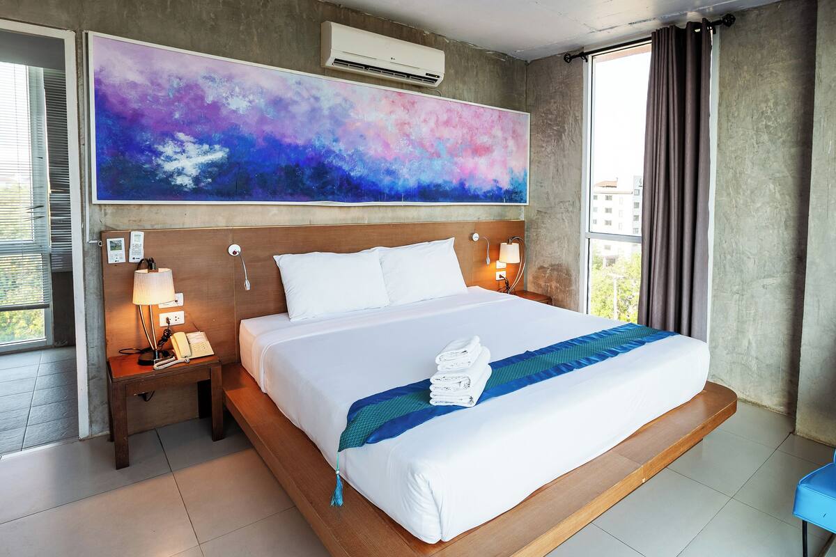 A hotel room with a large bed, a nightstand with a lamp, a blue chair, and a large abstract painting on the wall.