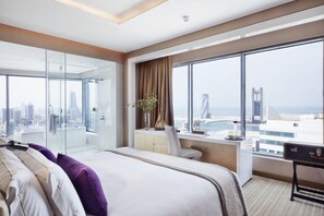 Una habitación de hotel moderna con una cama grande, un escritorio y vistas a un paisaje urbano a través de ventanas amplias.