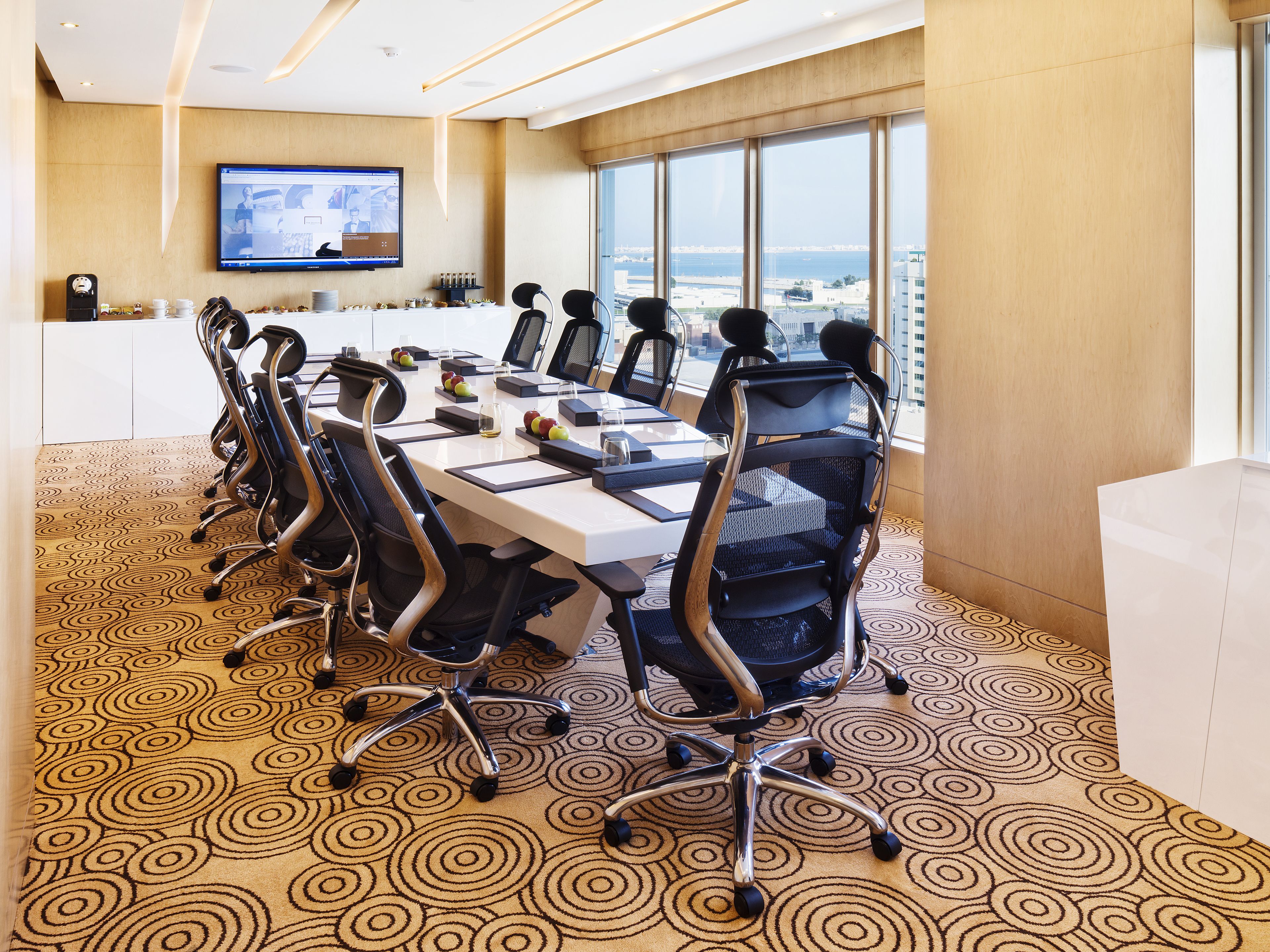 A conference room with a long table, multiple chairs, a television screen, and large windows offering a view of the city.