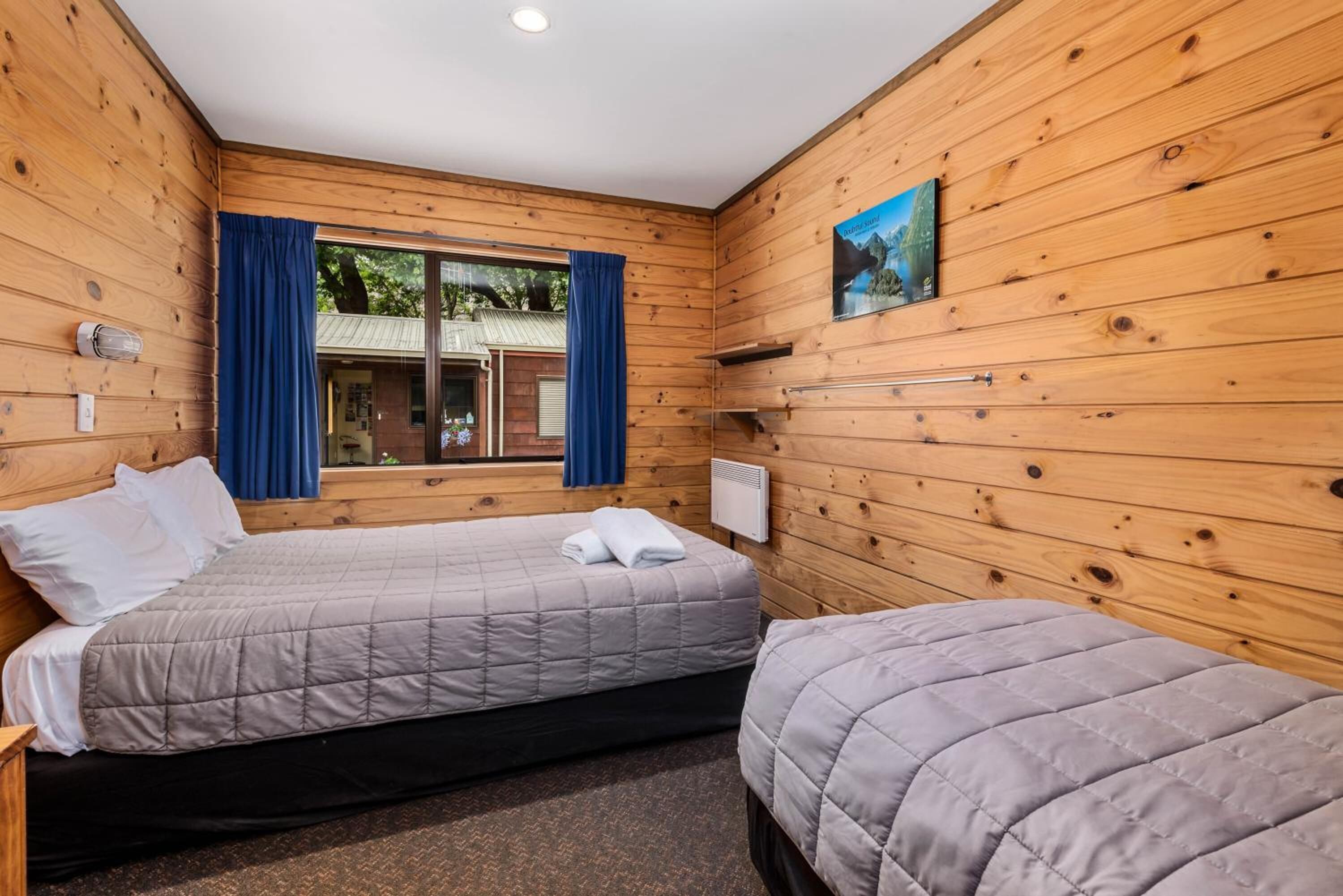 A wooden cabin room with two beds, a window with curtains, a radiator, and a painting on the wall.